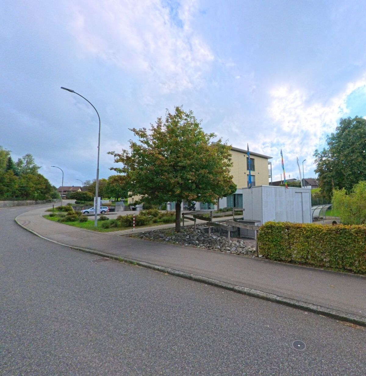 Vor einem modernen Verwaltungsgebäude mit wehenden Fahnen in Dürrenäsch (5724, Kanton Aargau) verläuft eine leicht gebogene Strasse mit Gehweg und Bäumen.