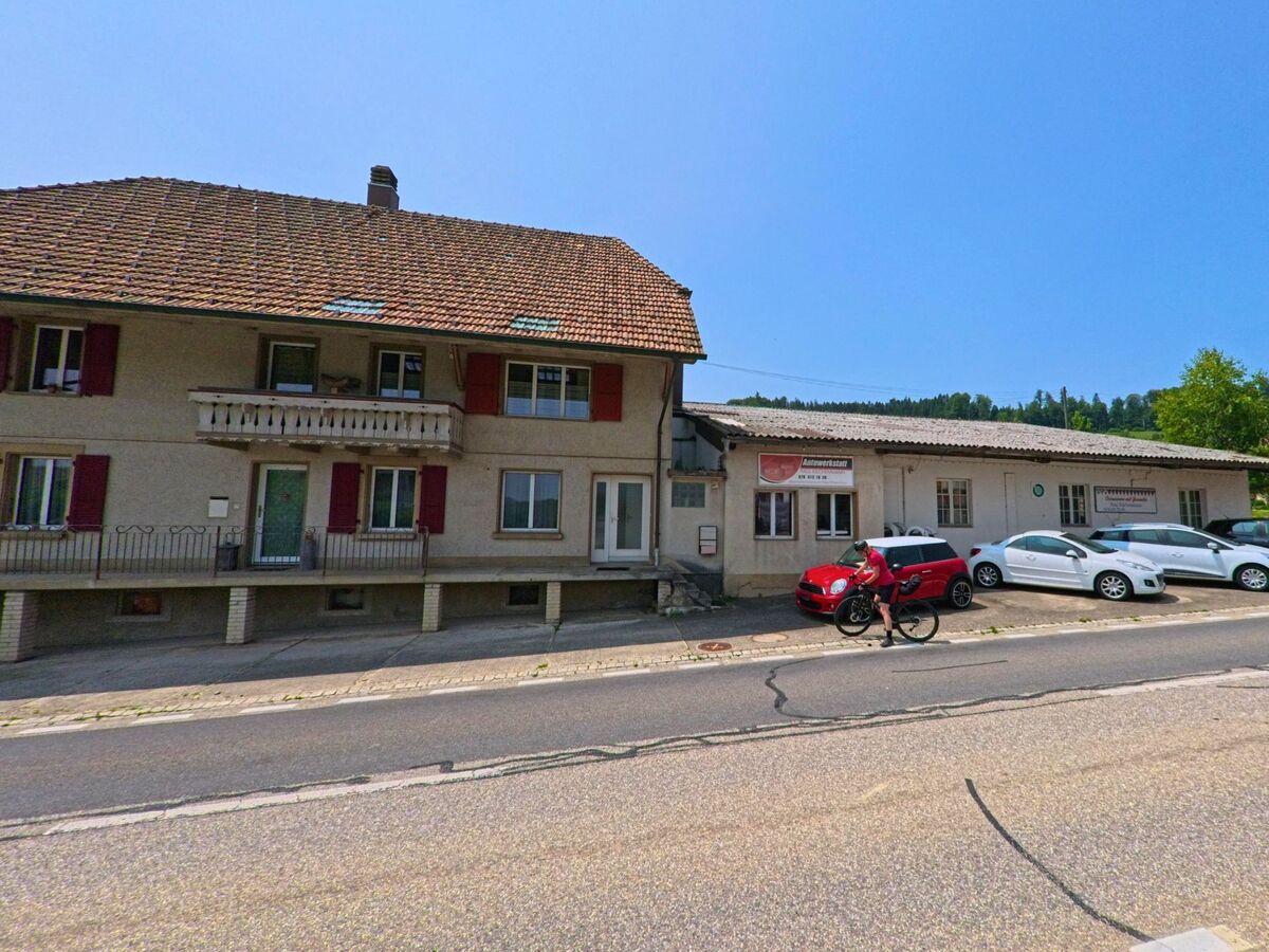 Entlang einer Straße in Dürrenroth (3465, Kanton Bern) befindet sich ein Wohnhaus mit angegliederter Autowerkstatt, vor der mehrere Fahrzeuge parken.
