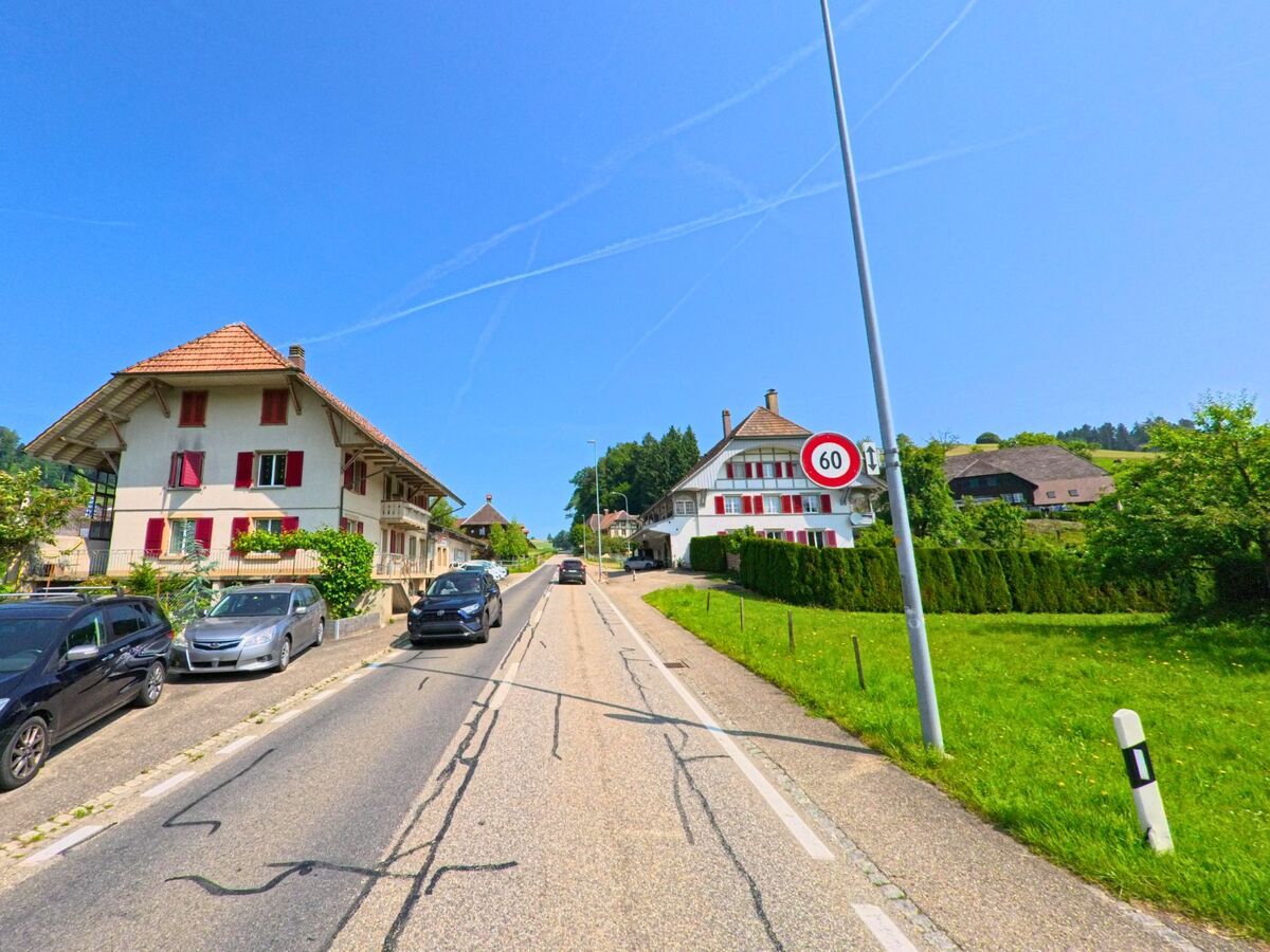 In Dürrenroth (3465, Kanton Bern) verläuft eine Dorfstraße mit traditionell gehaltenen Häusern und roten Fensterläden durch eine grüne, leicht hügelige Landschaft.