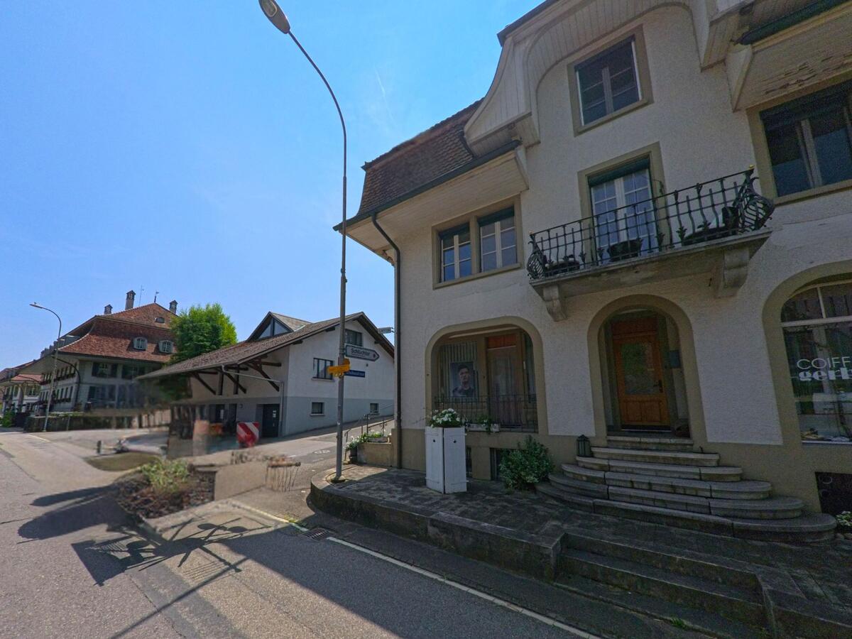 Im Ortszentrum von Dürrenroth (3465, Kanton Bern) liegt an einer Straßenkreuzung ein Gebäude mit Rundbogen-Eingang und Coiffeurgeschäft neben weiteren traditionellen Häusern