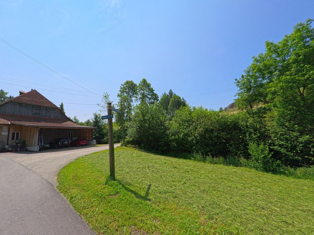 Am Rand eines Hofes in Dürrenroth (3465, Kanton Bern) zweigt ein Weg in ein bewaldetes Gebiet ab, markiert durch einen schlichten Holzwegweiser.