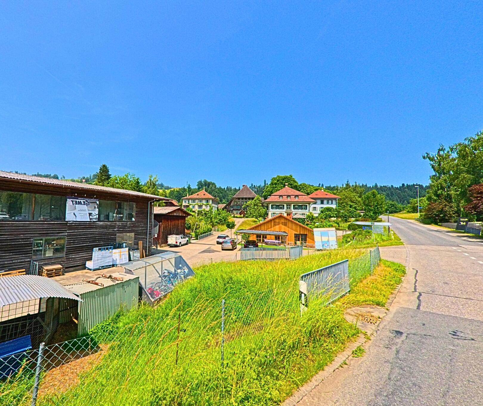 In Dürrenroth (3465, Kanton Bern) säumen Gewerbebauten und Wohnhäuser mit Walmdächern eine leicht ansteigende Landstrasse am Dorfrand.