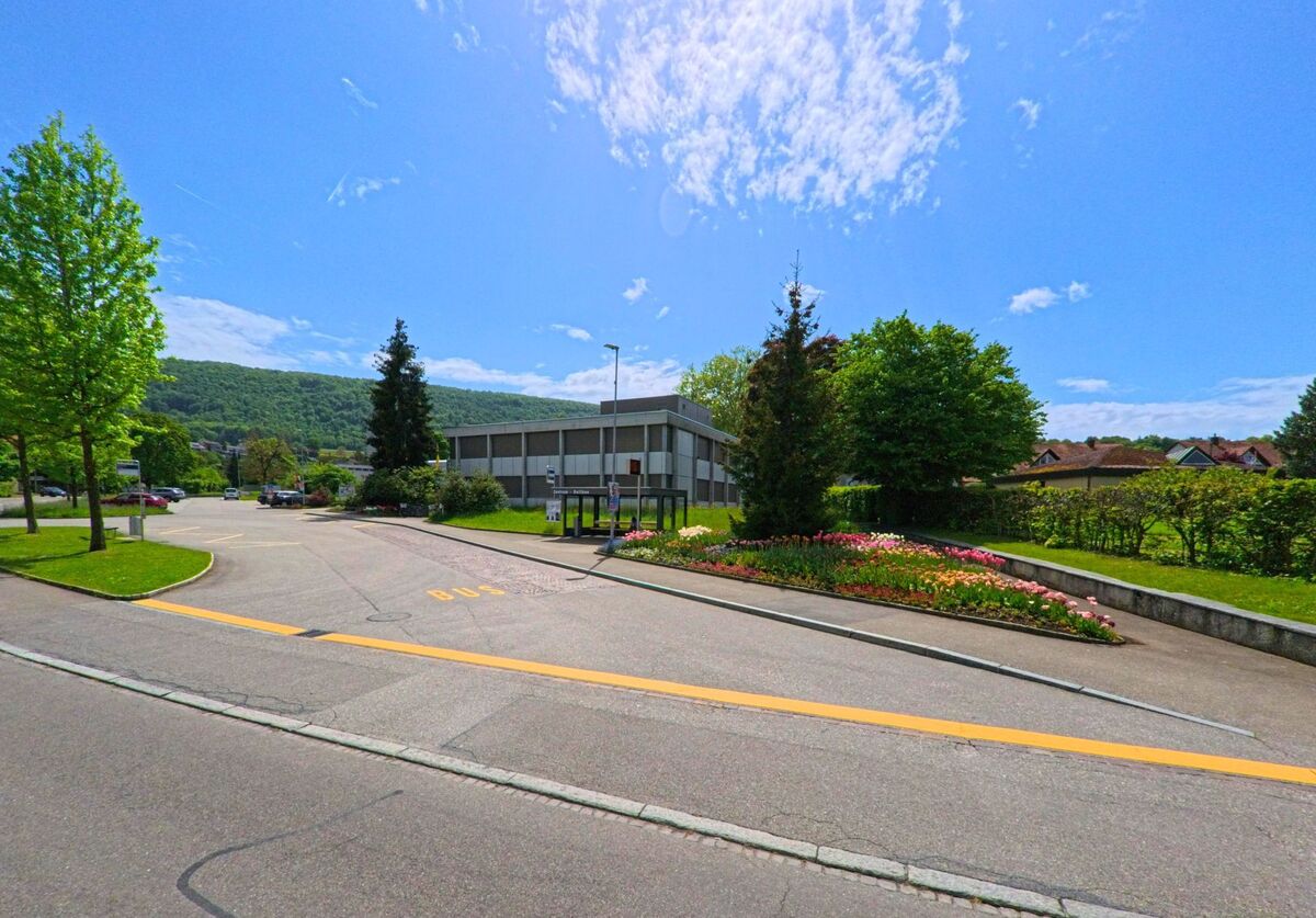 In Dulliken (4657, Kanton Solothurn) liegt eine Bushaltestelle neben einem schlichten Gebäude, umgeben von farbenfrohen Blumenbeeten und grüner Umgebung mit Hügelblick.