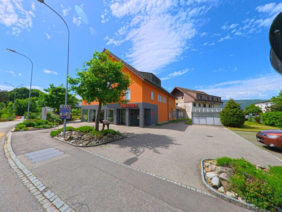 In Dulliken (4657, Kanton Solothurn) steht ein orangefarbenes Gebäude mit der Aufschrift „Raiffeisen“ an einer Kreuzung, umgeben von gepflegten Beeten und modernen Wohnhäusern.