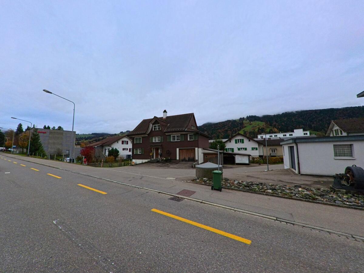 Ein braunes Wohnhaus mit gepflegtem Vorplatz steht in Ebnat-Kappel (9642, Kanton St. Gallen) an einer breiten Strasse, umgeben von weiteren Hausern und den bewaldeten Huegeln im Hintergrund.