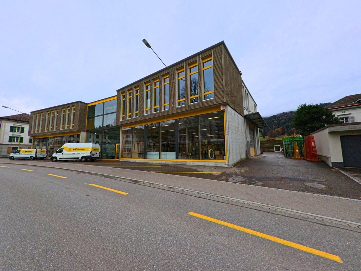 Ein Gewerbebau mit grossen Glasflaechen und gelben Akzenten praegt in Ebnat-Kappel (9642, Kanton St. Gallen) die Strasse, waehrend Lieferwagen davor parken und Huegel im Hintergrund liegen.