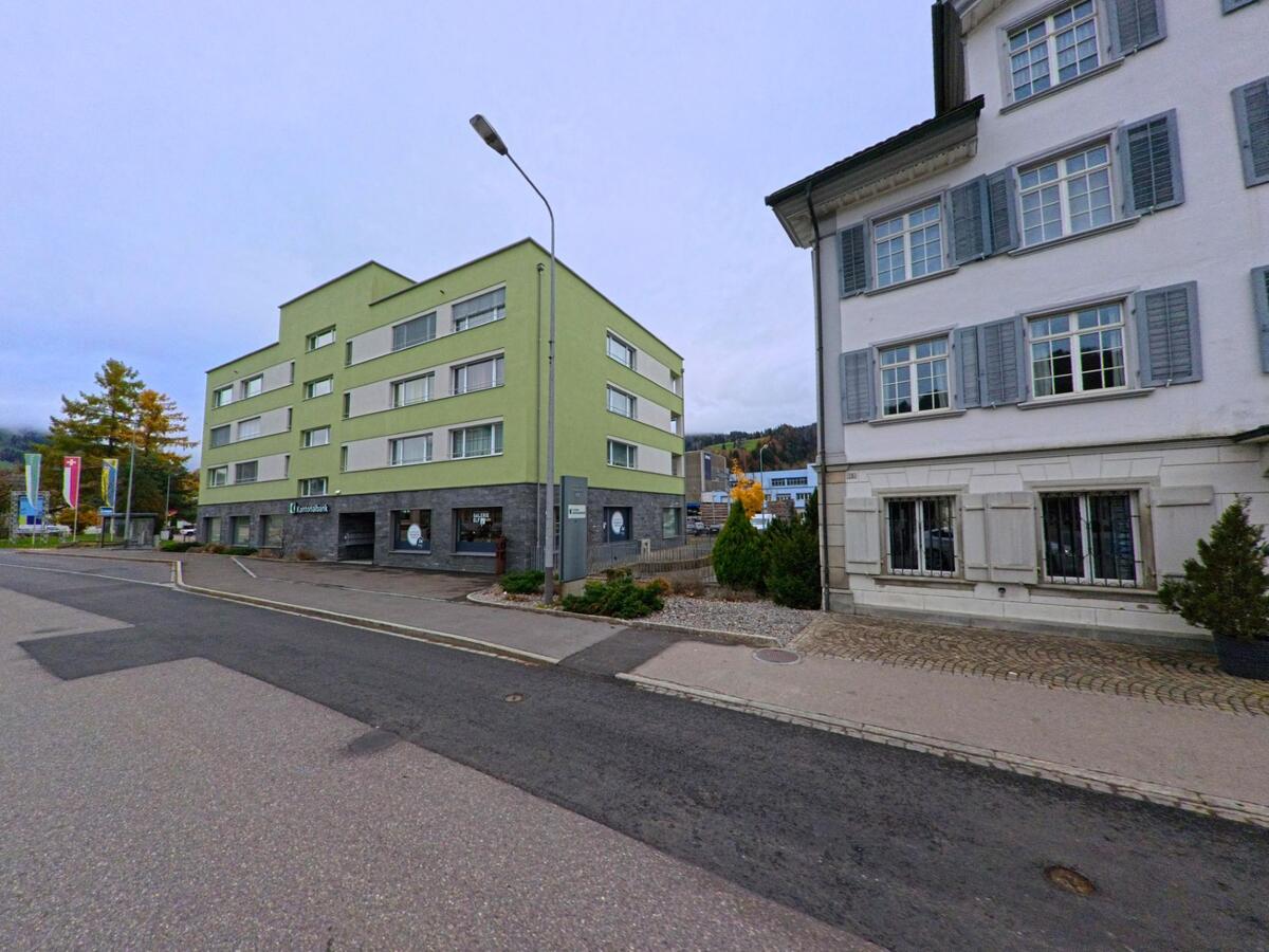 Ein gruenes Mehrfamilienhaus mit Gewerbeflaechen im Erdgeschoss steht in Ebnat-Kappel (9642, Kanton St. Gallen) an einer breiten Strasse, flankiert von weiteren Gebaeuden und herbstlichen Baumen.
