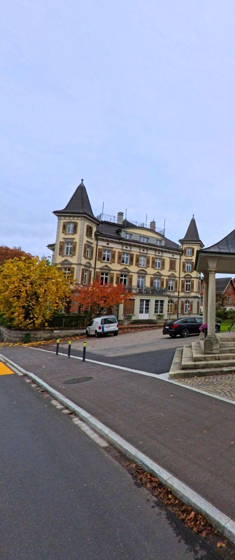 Ein herrschaftliches Gebaeude mit Tuermen und verzierter Fassade erhebt sich in Ebnat-Kappel (9642, Kanton St. Gallen) hinter herbstlich gefaerbten Baeumen und einer kleinen Platzflaeche.