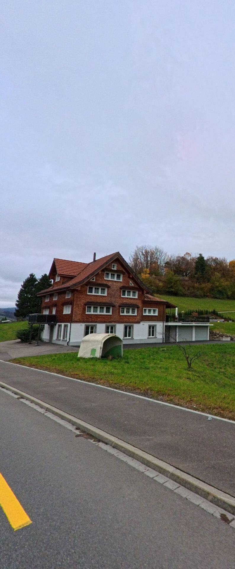 Ein traditionelles Holzhaus mit rotem Dach steht in Ebnat-Kappel (9642, Kanton St. Gallen) am Rand einer leicht ansteigenden Wiese unter herbstlich gefaerbten Baumen.
