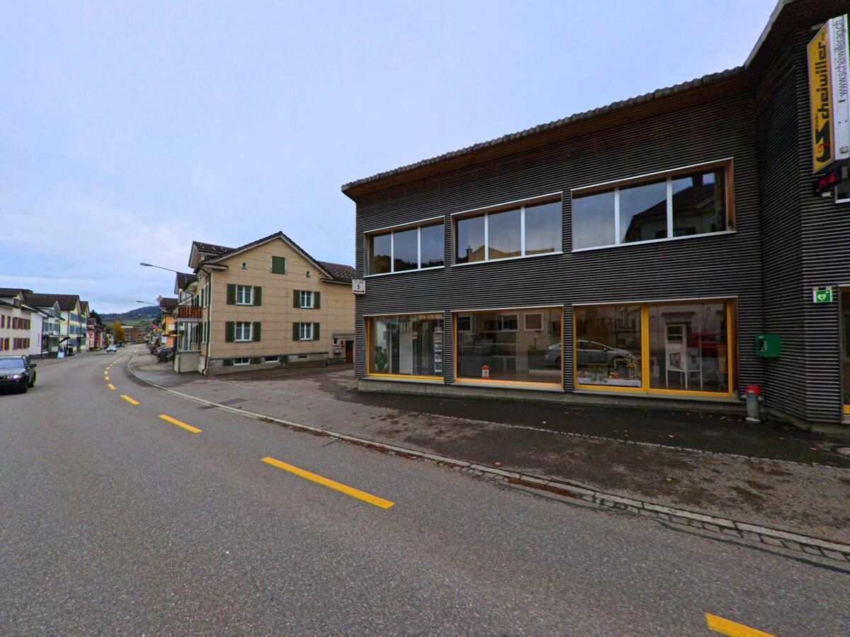 Ein modernes dunkles Gewerbegebaeude mit grossen Schaufenstern steht in Ebnat-Kappel (9642, Kanton St. Gallen) an einer kurvigen Strasse, umgeben von traditionellen Wohnhaeusern.