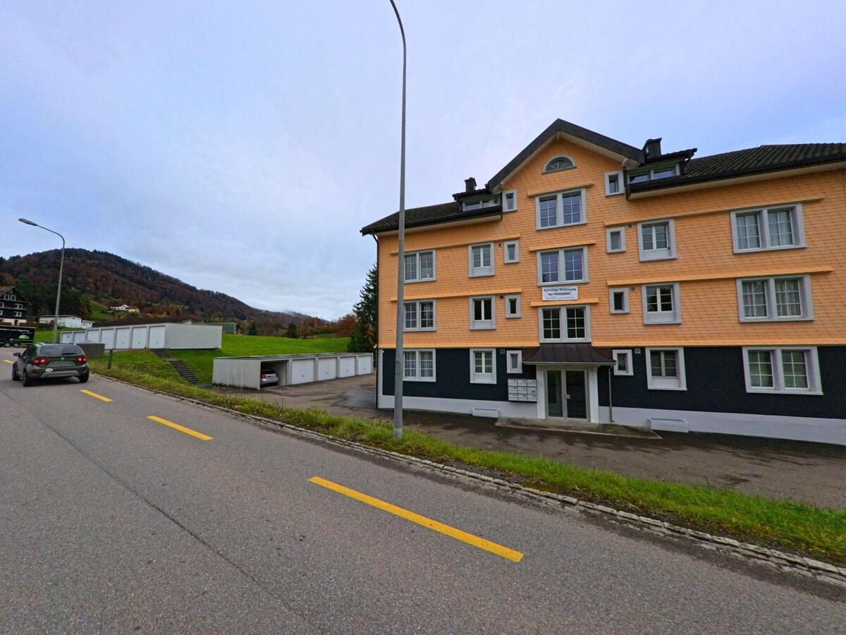 Ein modernes Mehrfamilienhaus mit apricotfarbener Fassade steht in Ebnat-Kappel (9642, Kanton St. Gallen) an einer leicht ansteigenden Strasse mit Blick auf die umliegenden Huegel.