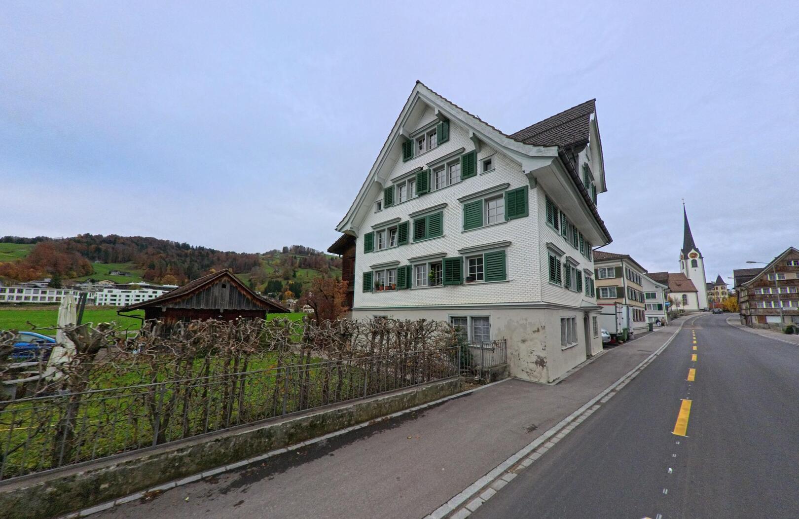 Ein traditionelles Wohnhaus mit gruene Fensterlaeden steht in Ebnat-Kappel (9642, Kanton St. Gallen) an einer leicht ansteigenden Strasse mit Blick auf die umliegenden Huegel.