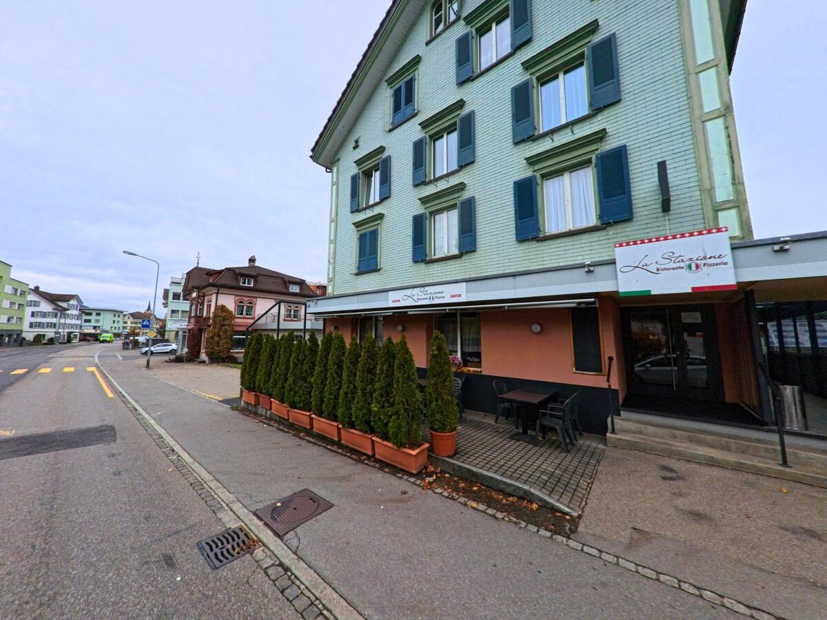 Ein gruenes Wohnhaus mit angeschlossenem Restaurant steht in Ebnat-Kappel (9642, Kanton St. Gallen) an einer breiten Strasse, waehrend weitere Gebaeude die Ortschaft praegen.