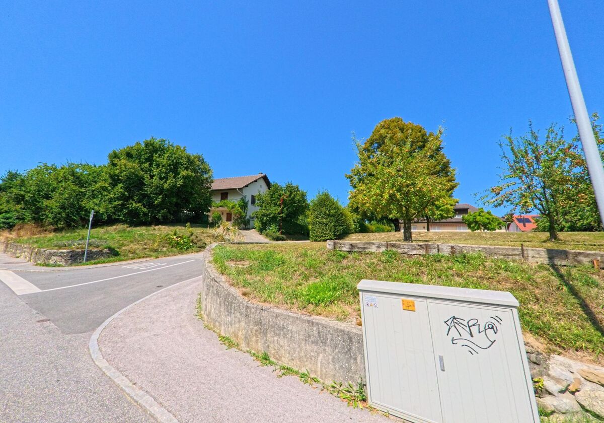 Ein Wohnhaus auf einer leichten Anhöhe in Echichens (1112, Kanton Waadt) ist von Bäumen und Gärten umgeben, davor verläuft eine schmale Strasse.