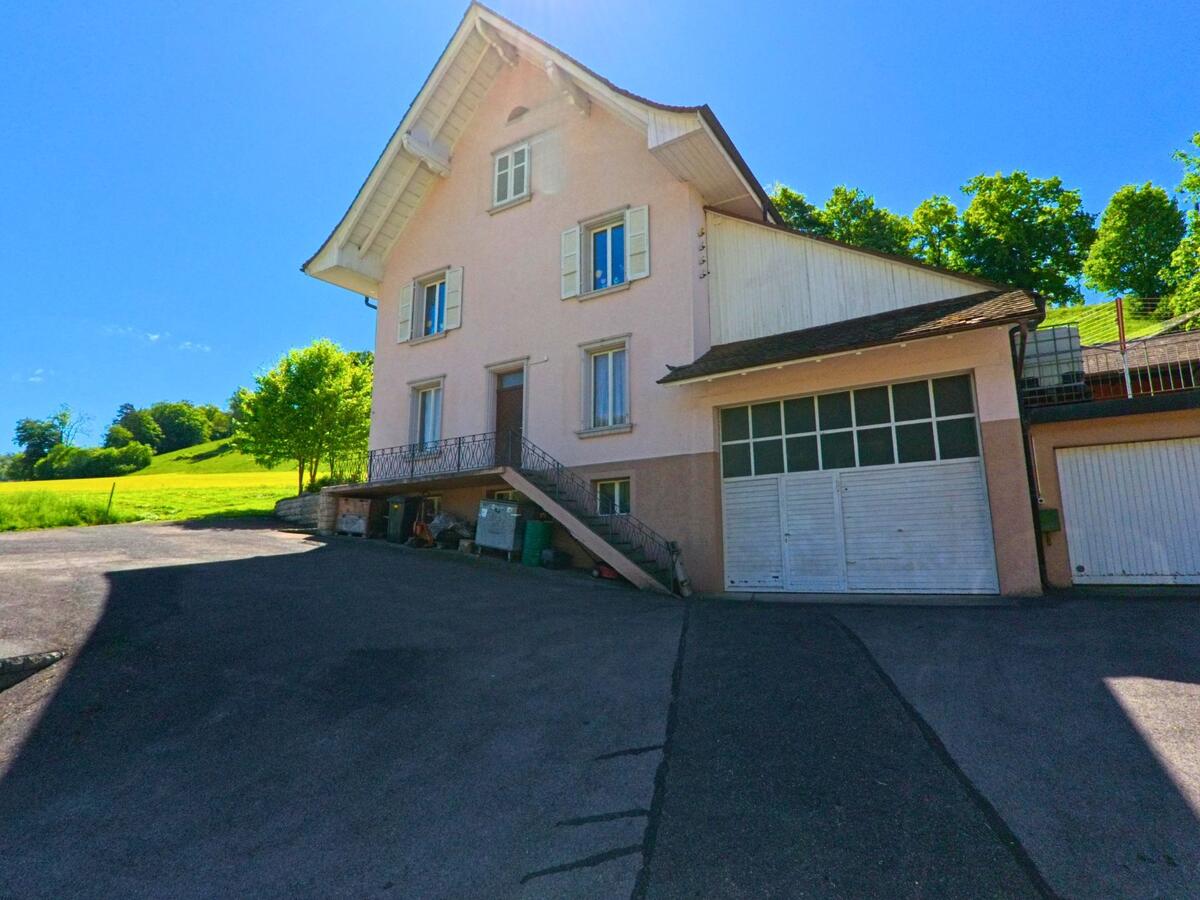 In Ederswiler (2813, Kanton Jura) führt die Straße um die Ecke eines weißen Gebäudes mit einer markanten Kirchturmuhr. Im Hintergrund sind weitere Gebäude und sanfte Hügel sichtbar.