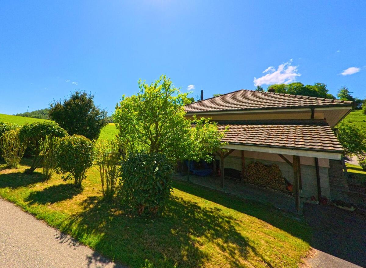 In Ederswiler (2813, Kanton Jura) ist ein kleines Haus mit einer überdachten Terrasse zu sehen, umgeben von grünen Sträuchern und Wiese. Der sonnige Himmel und die sanften Hügel im Hintergrund verleihen der Szene eine ländliche Atmosphäre.
