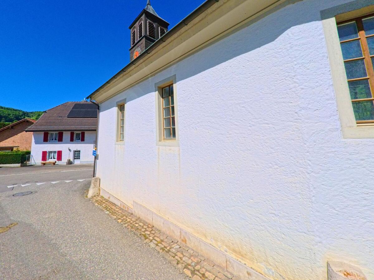 In Ederswiler (2813, Kanton Jura) ist ein großes, pinkfarbenes Gebäude mit einem Garagenbereich und einem Eingang auf der Seite zu sehen. Der umliegende Bereich ist von grünen Wiesen und Bäumen geprägt, und der klare Himmel ergänzt die ländliche Atmosphäre.