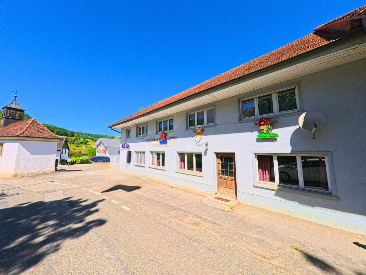 In Ederswiler (2813, Kanton Jura) ist ein Gebäude mit einem Restaurant-Schild und bunten Kinderfiguren an der Fassade zu sehen. Ein graues Auto parkt vor dem Gebäude, während im Hintergrund eine Kirche und grüne Hügel sichtbar sind.