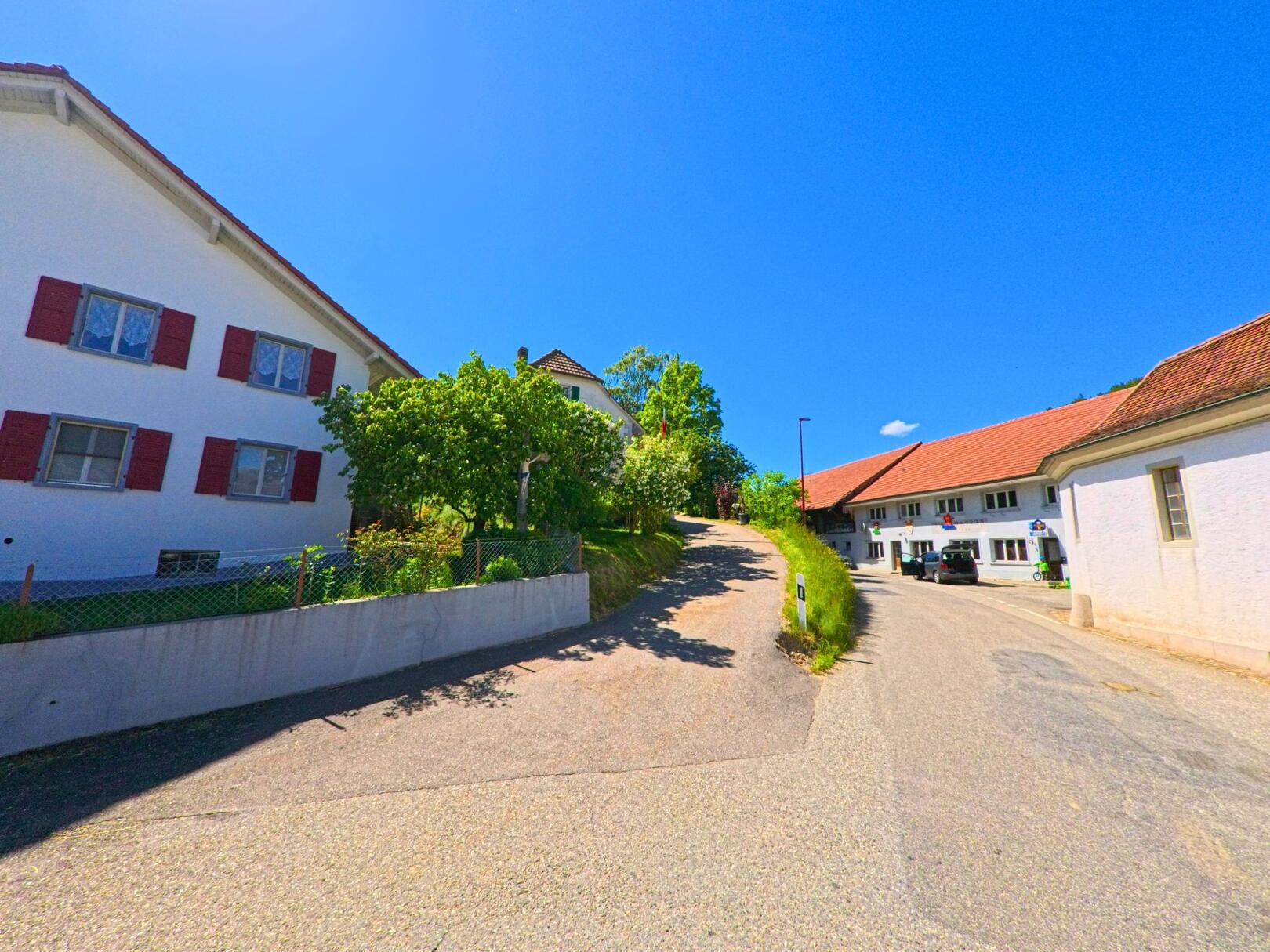 In Ederswiler (2813, Kanton Jura) ist eine weisse Wohnhausfassade mit roten Fensterläden an einer Strassenkurve zu sehen. Ein schwarzes Auto steht am Strassenrand, und im Hintergrund sind weitere Gebäude sowie grüne Hügel sichtbar.