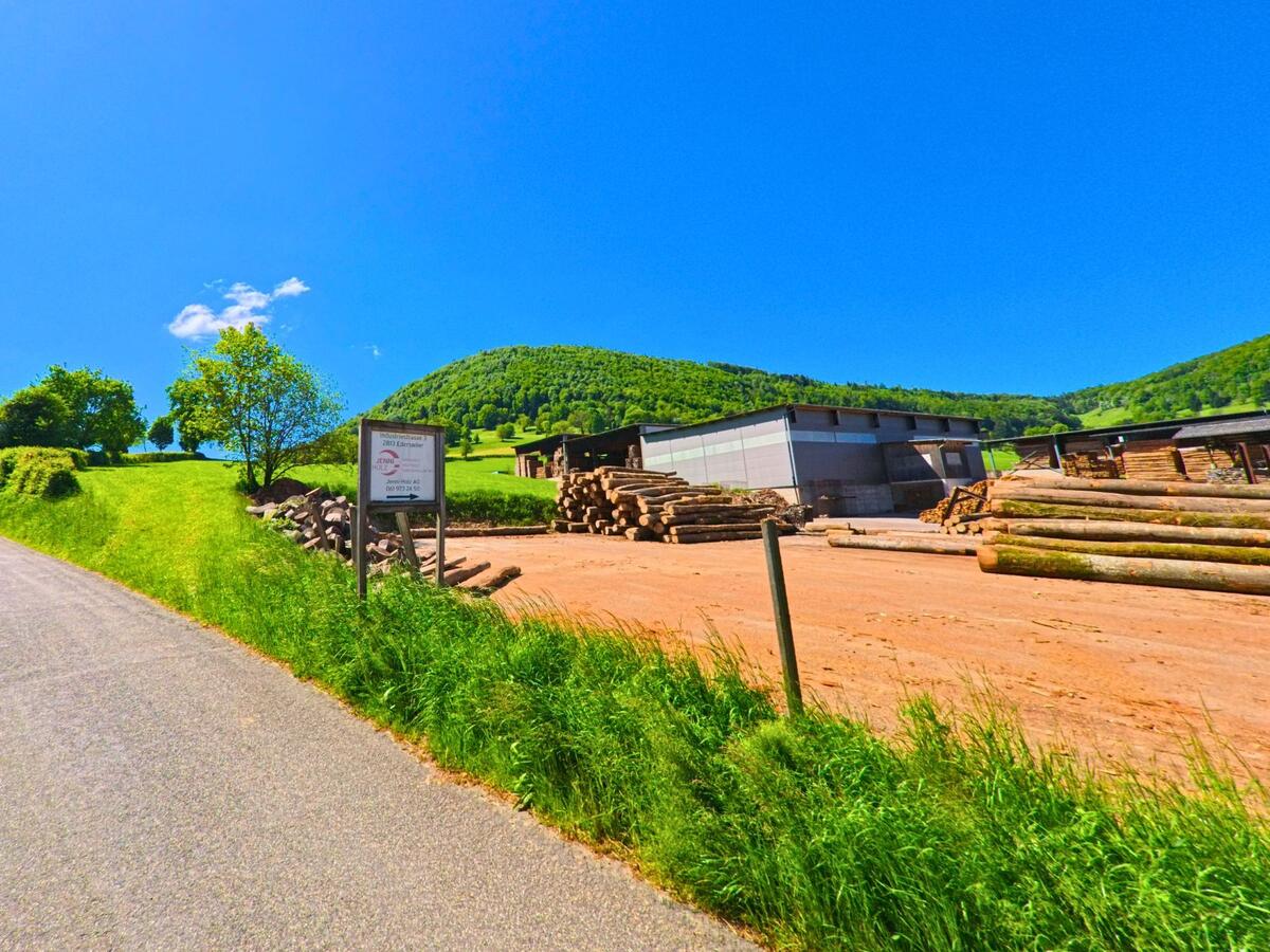 In Ederswiler (2813, Kanton Jura) zeigt sich ein Waldwirtschaftsbetrieb mit gestapelten Holzstämmen entlang einer Straße. Im Hintergrund erstreckt sich eine hügelige Landschaft mit grünen Wiesen und Bäumen.