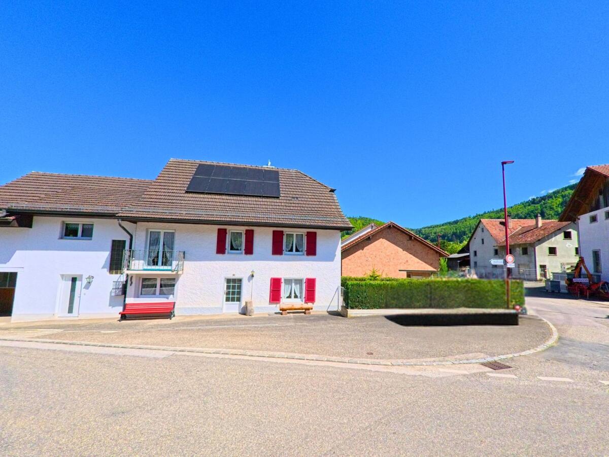 In Ederswiler (2813, Kanton Jura) ist eine weiße Wohnhausfassade mit roten Fensterläden an einer Strassenkurve zu sehen. Ein schwarzes Auto steht am Straßenrand, und im Hintergrund sind weitere Gebäude sowie grüne Hügel sichtbar.