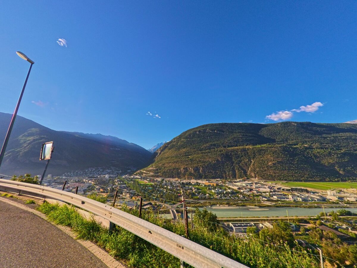 Von einer Strasse in Eggerberg (3939, Kanton Wallis) oeffnet sich der Blick ueber das Rhonetal mit der Stadt im Talgrund und den gruenen Haengen des Berges gegenüber.