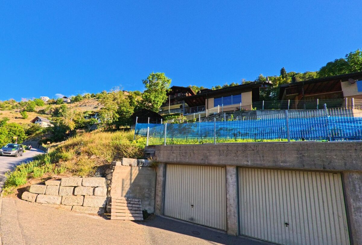 In Eggerberg (3939, Kanton Wallis) sind mehrere Wohnhaeuser am Hang gebaut, vor denen eine Reihe von Garagen und eine steile Strasse verlaufen.