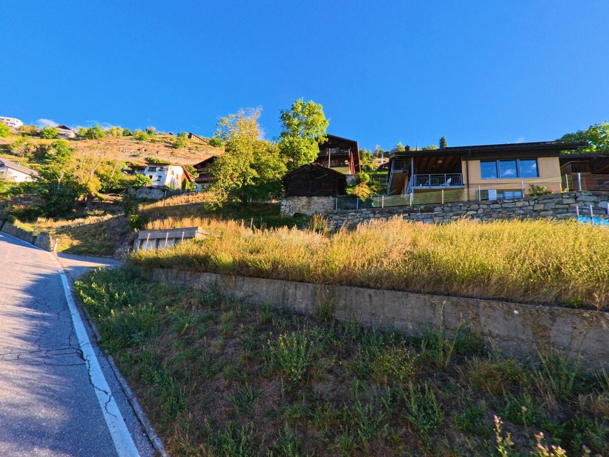 Wohnhaeuser in Eggerberg (3939, Kanton Wallis) reihen sich am Hang oberhalb einer steilen Strasse, umgeben von Gruenflaechen und Baumen.