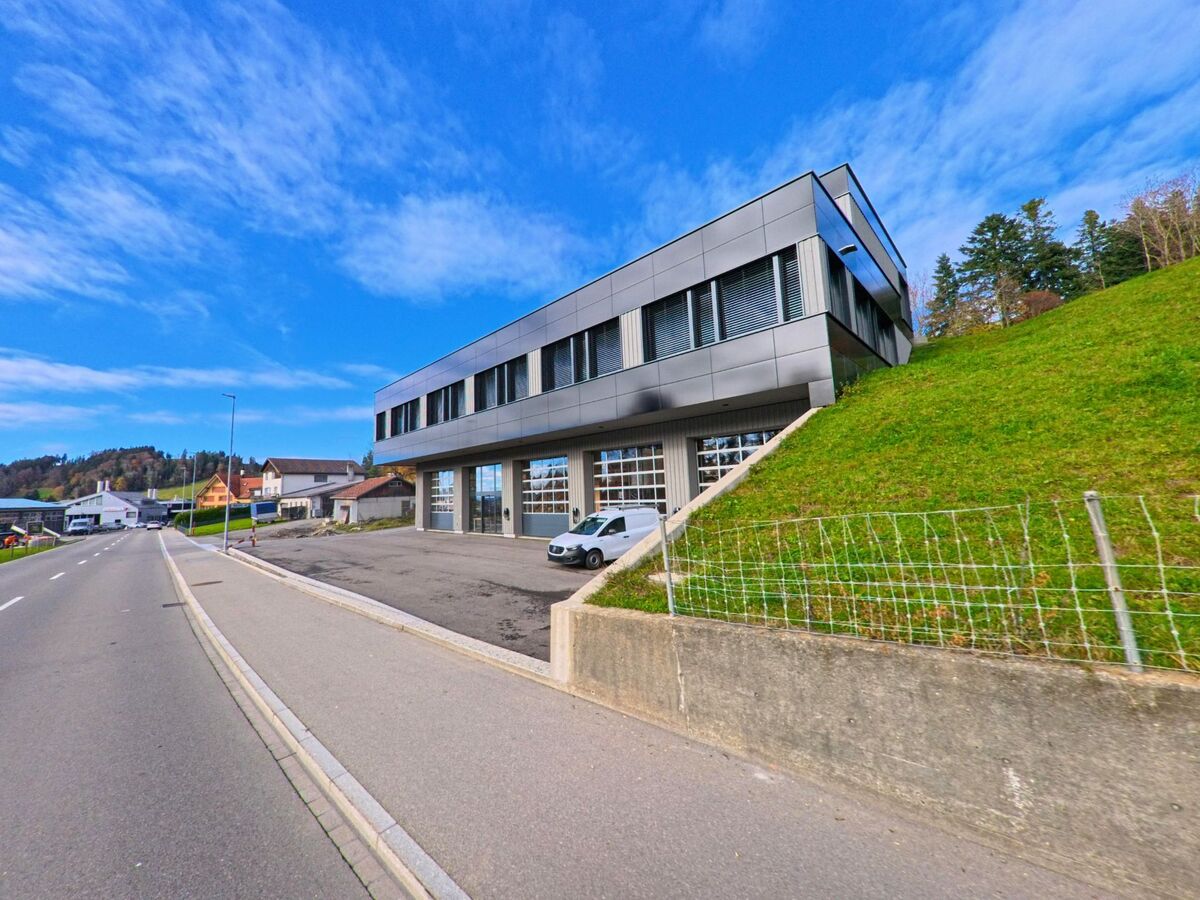 Ein modernes Gewerbegebaeude mit grossen Toren steht in Eggersriet (9036, Kanton St. Gallen) am Fuss eines gruenen Hangs direkt an der Durchgangsstrasse.