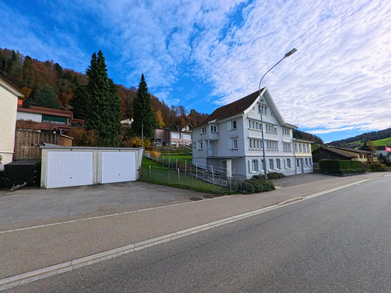 Ein traditionelles Wohnhaus steht in Eggersriet (9036, Kanton St. Gallen) an einer ruhigen Strasse, flankiert von Garagen und leicht ansteigendem Hang.