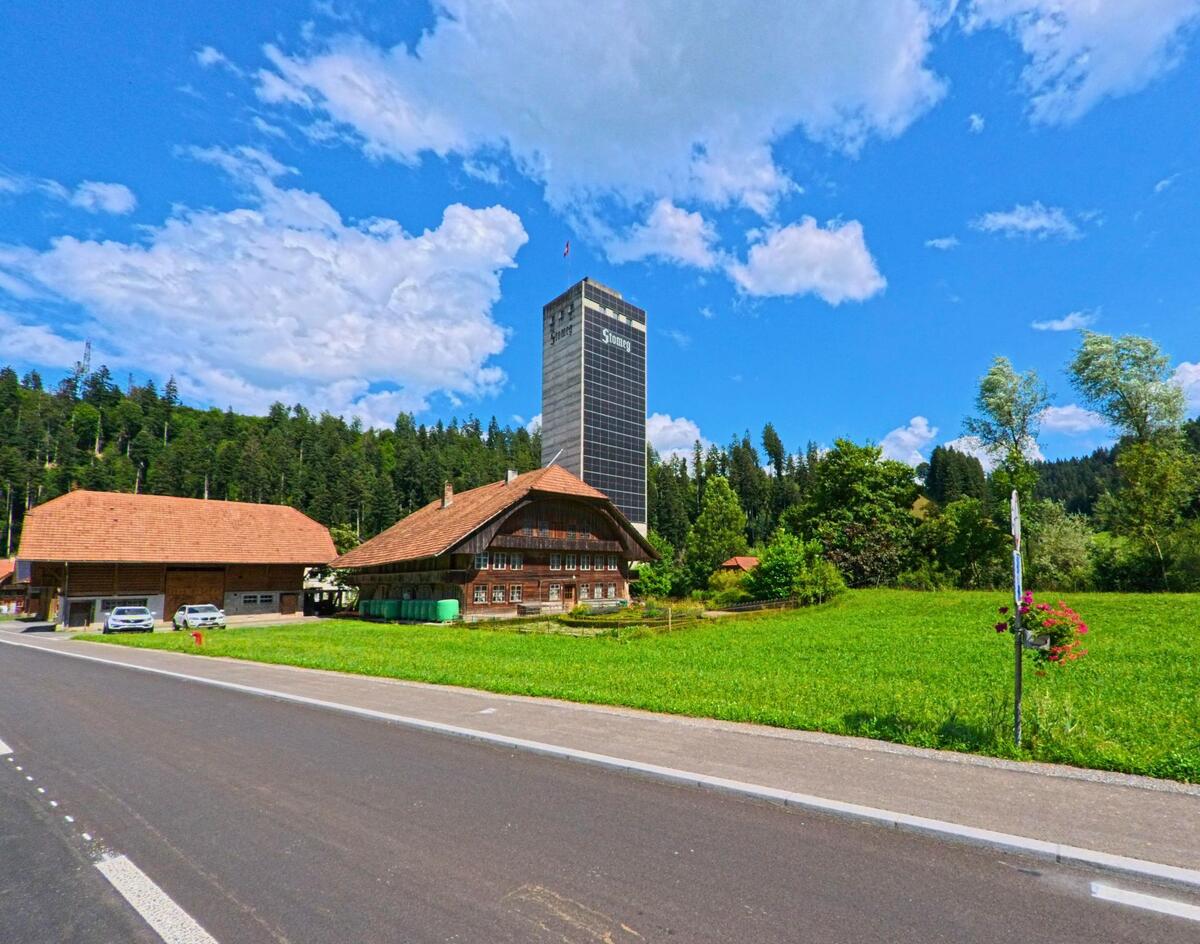 In Eggiwil (3537, Kanton Bern) steht ein traditionelles Bauernhaus mit grossen Dachflaechen vor einem modernen Hochhaus der Firma Simona, eingerahmt von Wiesen und Wald.