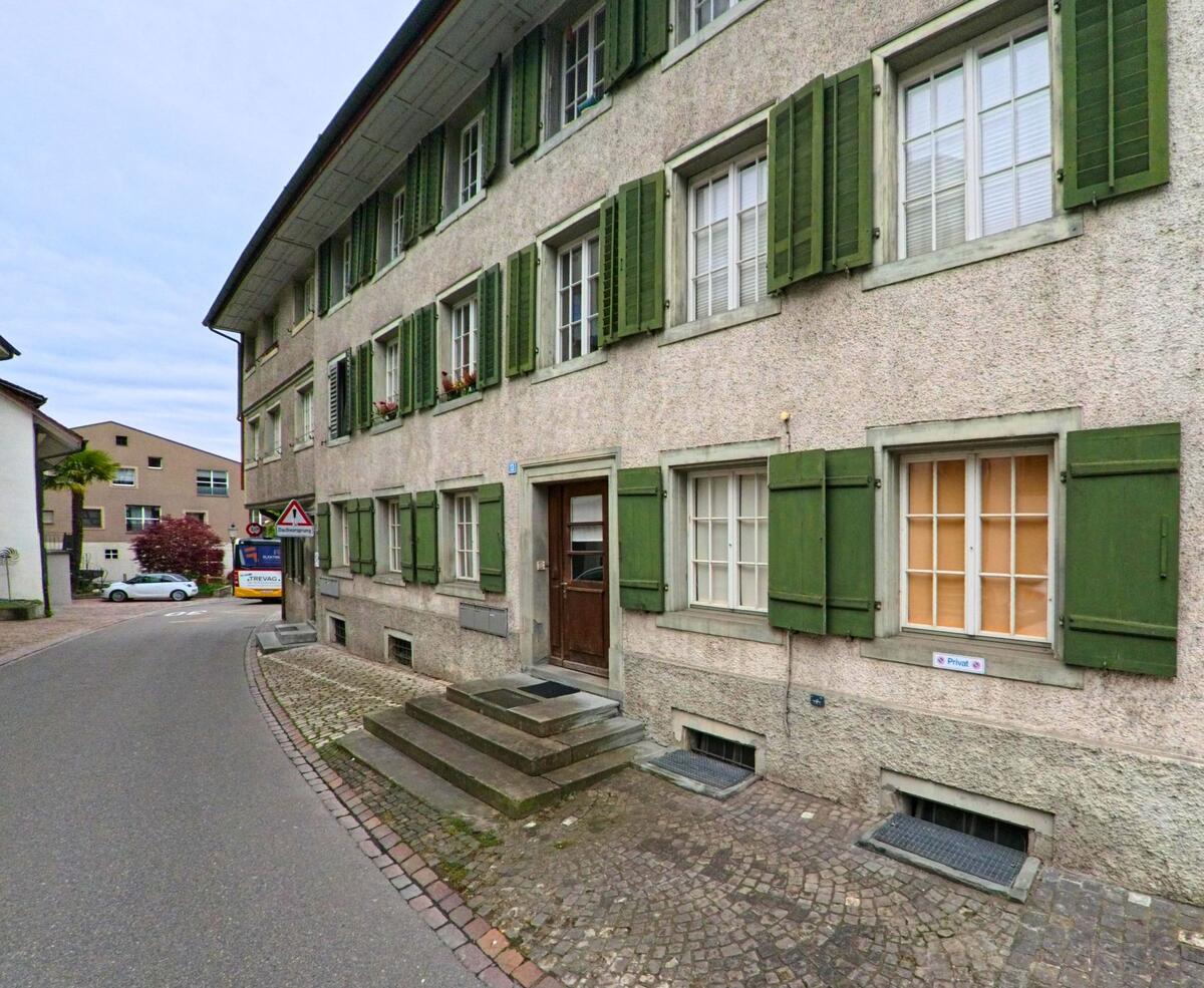  In einer engen Gasse von Eglisau, PLZ 8193 im Kanton Zürich, steht ein mehrstöckiges Haus mit grünen Fensterläden und einem Hauseingang mit Treppe.
