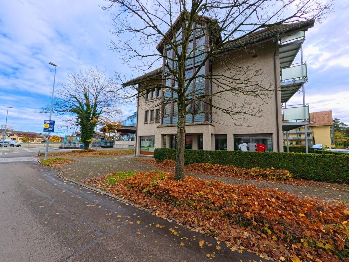 An einer Strassenecke in Egnach (9322, Kanton Thurgau) steht ein Gebäude mit grossen Fenstern und Balkonen, davor liegen braune Herbstblätter entlang des Gehwegs.