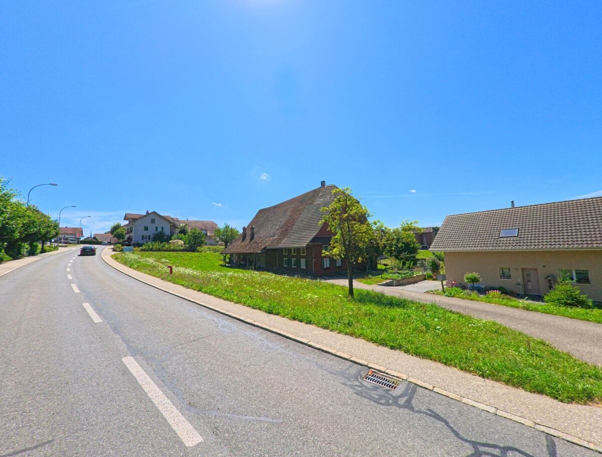 Entlang einer leicht ansteigenden Strasse in Egolzwil (PLZ 6243, Kanton Luzern) stehen traditionelle Wohnhaeuser mit Gaerten unter blauem Himmel.