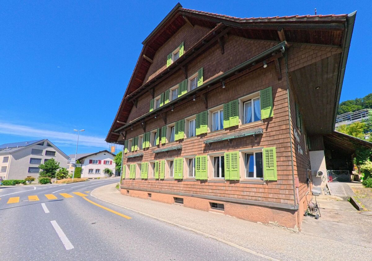 In Egolzwil (PLZ 6243, Kanton Luzern) steht ein traditionelles Holzhaus mit gruenen Fensterlaeden an einer Kurve mit Fussgaengerstreifen und moderner Wohnbebauung im Hintergrund.