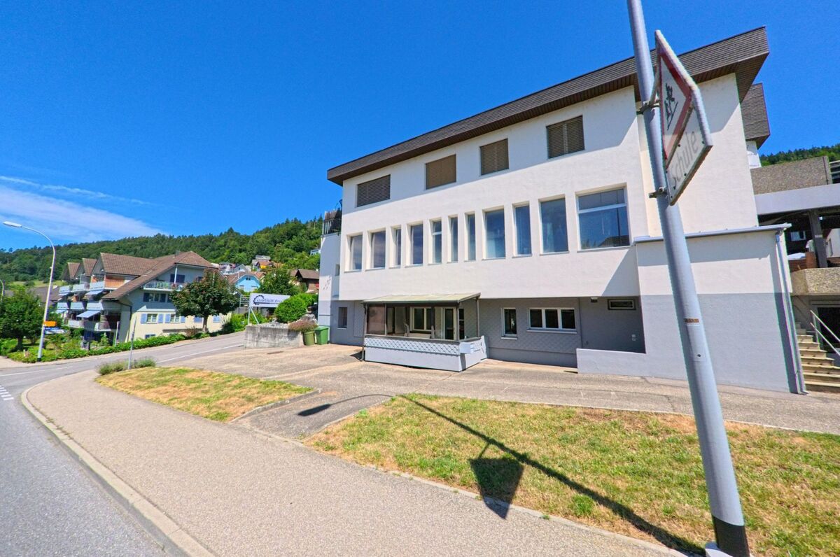 In Egolzwil (PLZ 6243, Kanton Luzern) steht ein weiss gestrichenes Schulgebaeude an einer leicht ansteigenden Strasse mit Wohnhaeusern und bewaldetem Hang im Hintergrund.