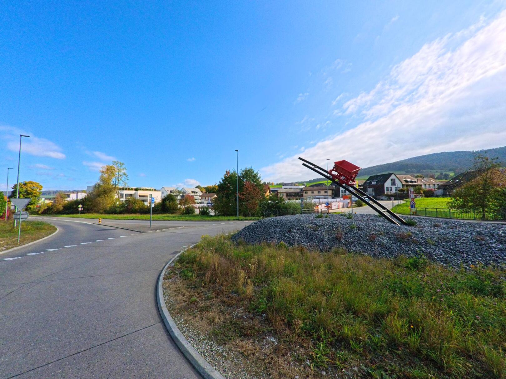 Am Kreisel in Ehrendingen (5420, Kanton Aargau) steht eine moderne Skulptur mit einer roten Lorenmulde auf schrägen Schienen, umgeben von Wohnhäusern und sanften Hügeln im Hintergrund.