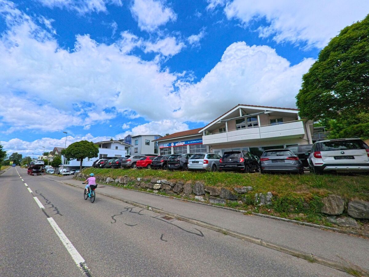 In Eich (6205, Kanton Luzern) verläuft eine Hauptstrasse an einem Autohaus mit grossem Parkplatz vorbei, waehrend ein Kind auf dem Velo den Gehweg entlangfaehrt.