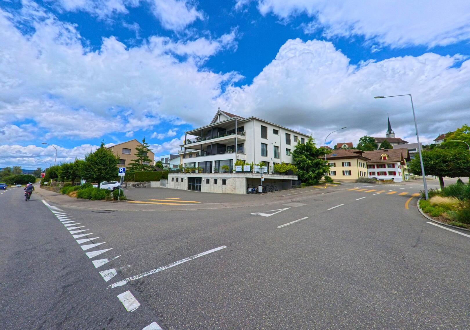 In Eich (6205, Kanton Luzern) steht ein modernes Wohnhaus mit Balkonen an einer Strassenkreuzung, im Hintergrund sind traditionelle Gebaeude und der Kirchturm des Dorfes zu erkennen