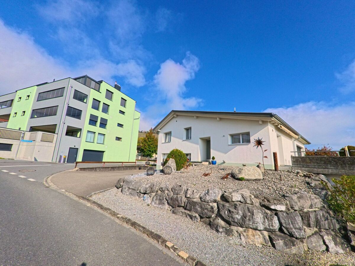 In Eichberg (9453, Kanton St. Gallen) stehen ein modernes Mehrfamilienhaus und ein niedriges weisses Wohnhaus an einer leicht gekruemmten Strasse mit steiniger Gartenanlage.