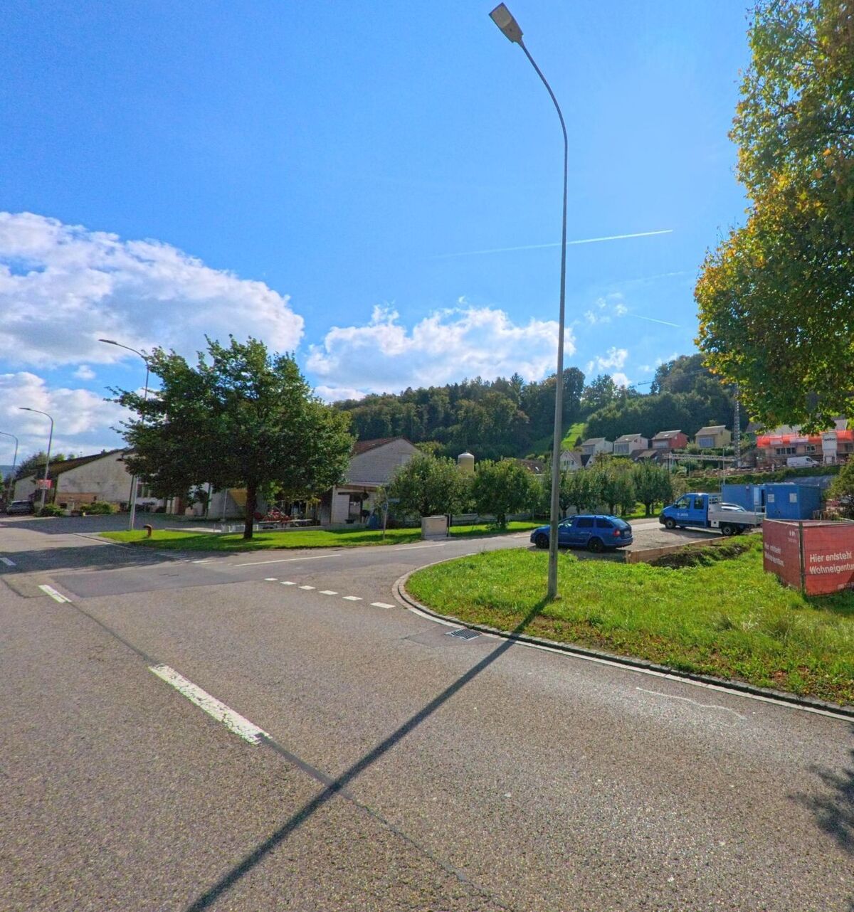 Am Ortseingang von Eiken (5074, Kanton Aargau) führt eine Strasse mit Neubauten und Bäumen entlang eines Hügels mit Blick auf Wohnsiedlungen und Obstgärten.