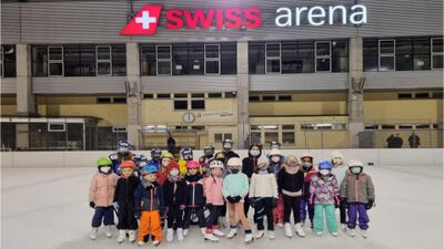 In Kloten (8302, Kanton Zuerich) steht eine grosse Gruppe von Kindern mit Helmen und Schlittschuhen auf dem Eis der Swiss Arena und posiert fuer ein gemeinsames Foto.