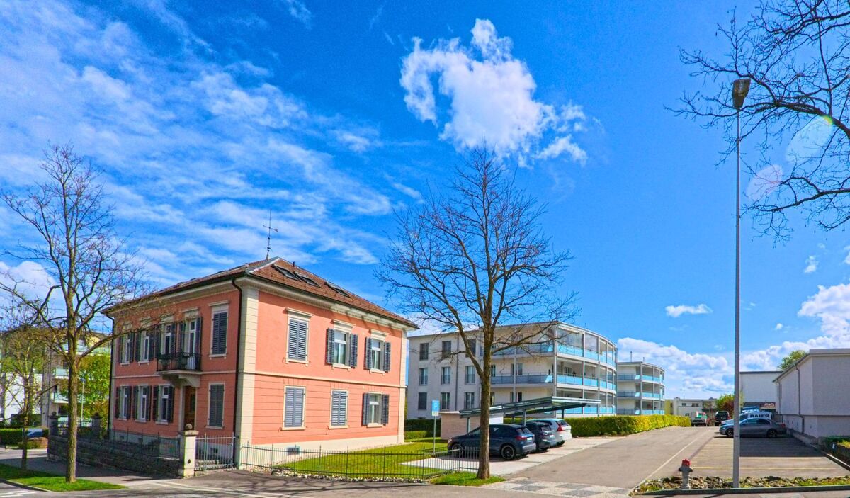  In Elgg, PLZ 8353 im Kanton Zürich, steht eine historische Villa mit rosafarbener Fassade im Kontrast zu modernen Wohnbauten im Hintergrund.