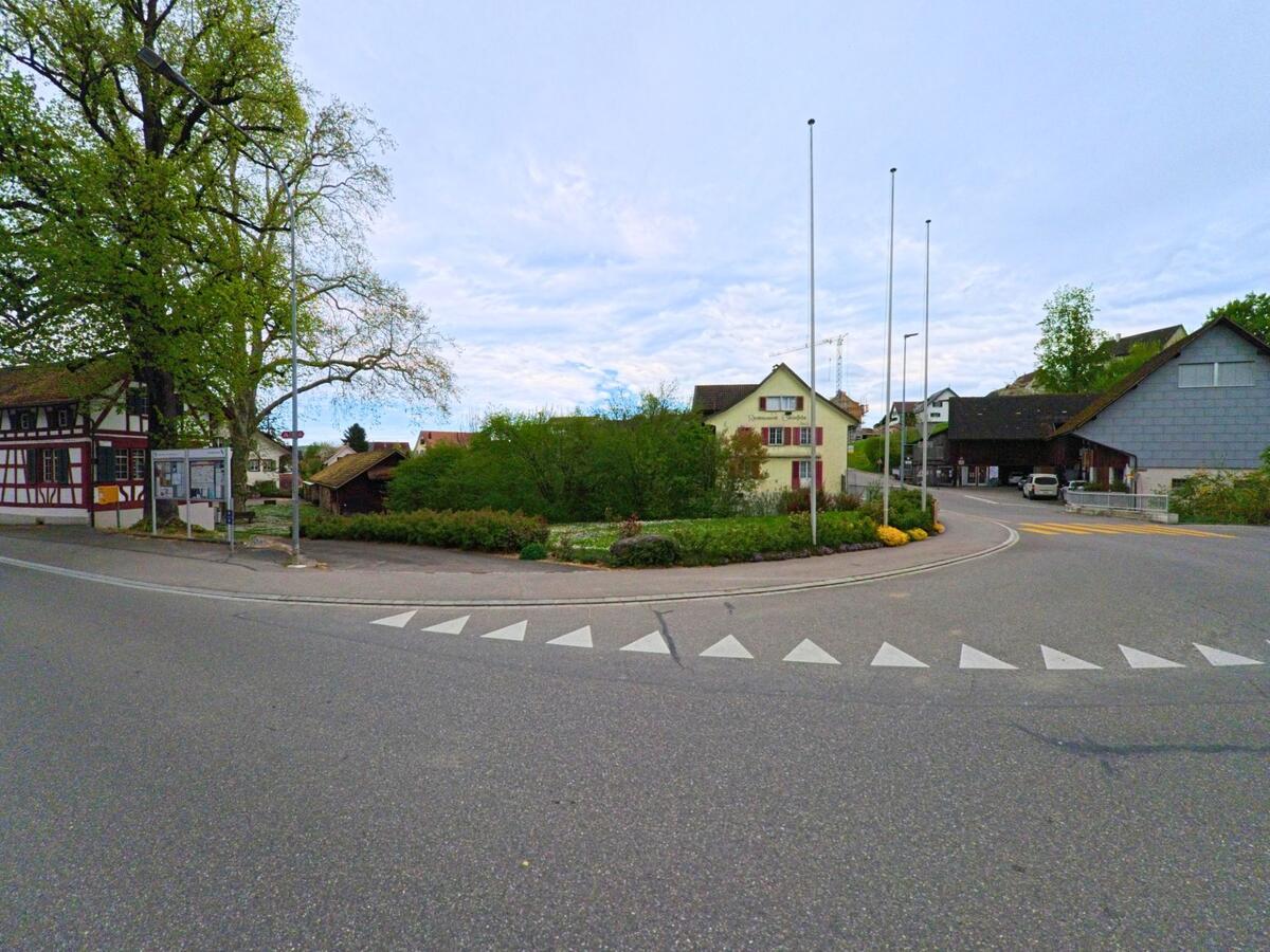 Im Zentrum von Ellikon an der Thur, PLZ 8548 im Kanton Zürich, stehen traditionelle Wohnhäuser und ein öffentliches Anschlagbrett an einer kleinen Platzanlage mit Fahnenmasten.