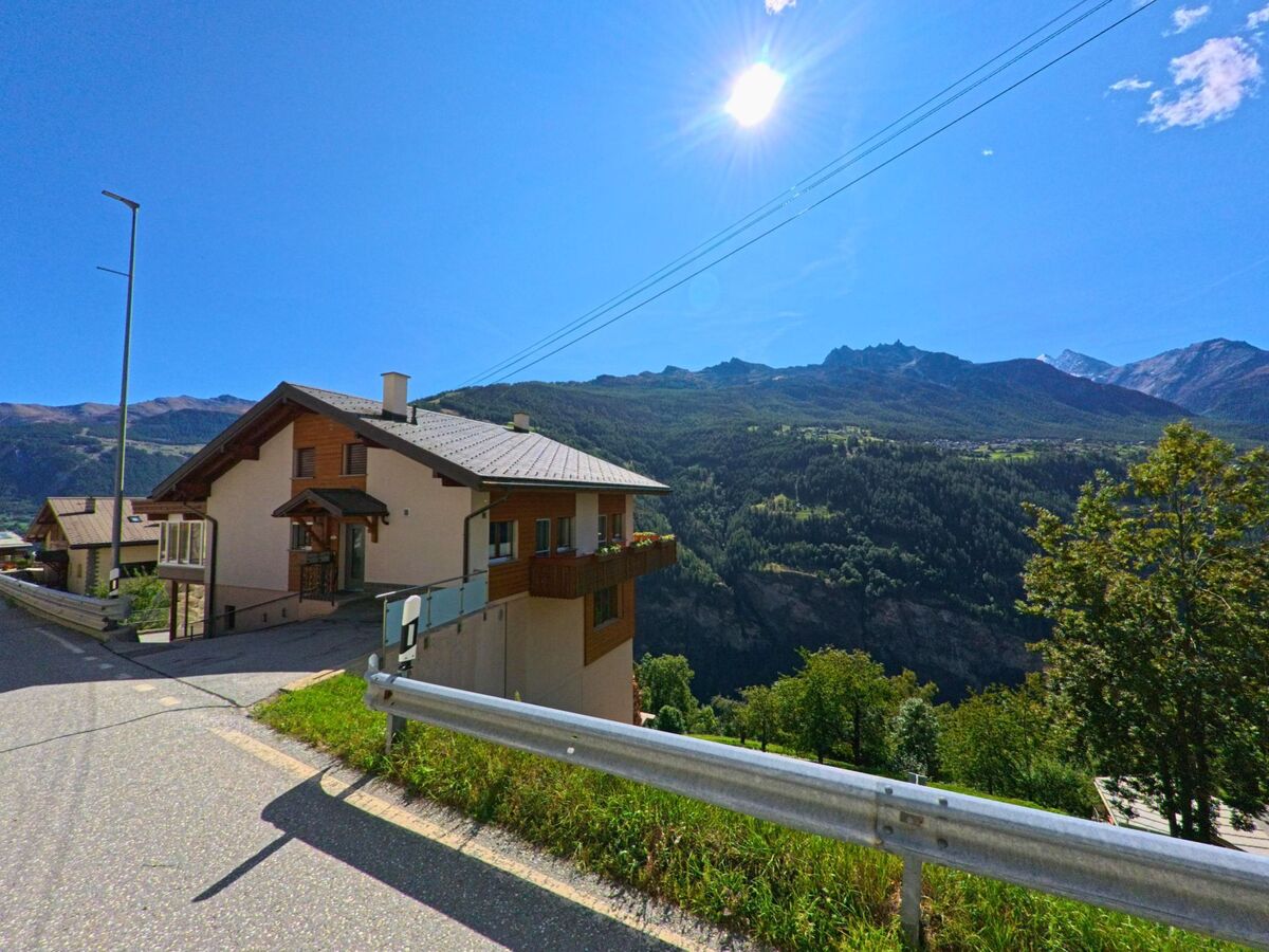 Ein Einfamilienhaus in Embd (3926, Kanton Wallis) steht an einer Hangstrasse mit Blick auf bewaldete Berghaenge und Gipfel im Hintergrund.