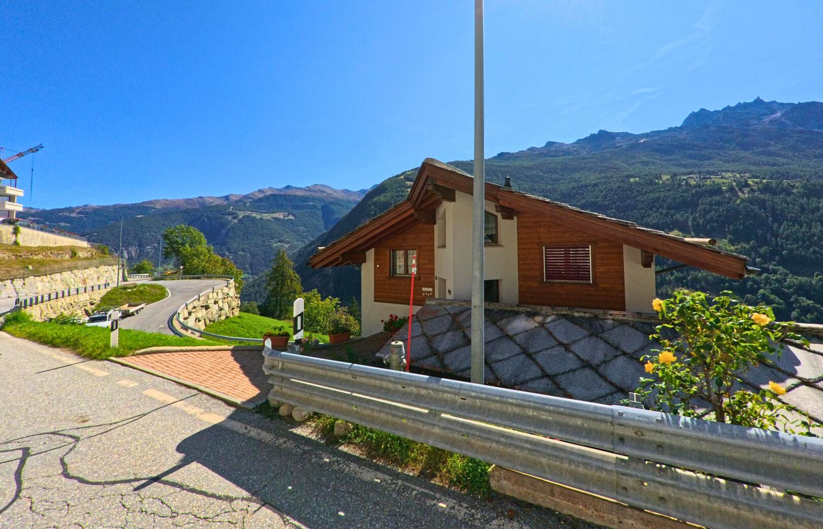 Ein Wohnhaus mit Holzelementen steht am Hang in Embd (3926, Kanton Wallis), daneben verläuft eine kurvenreiche Bergstrasse mit Blick auf die umliegenden Berge.
