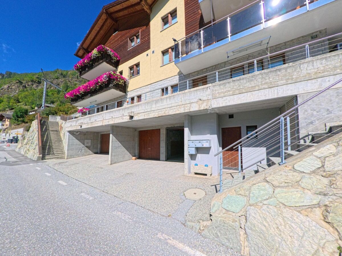 Ein Wohnhaus in Embd (3926, Kanton Wallis) zeigt Balkone mit leuchtend pinken Blumenkasten, darunter befinden sich Garagen und Hauseingaenge.