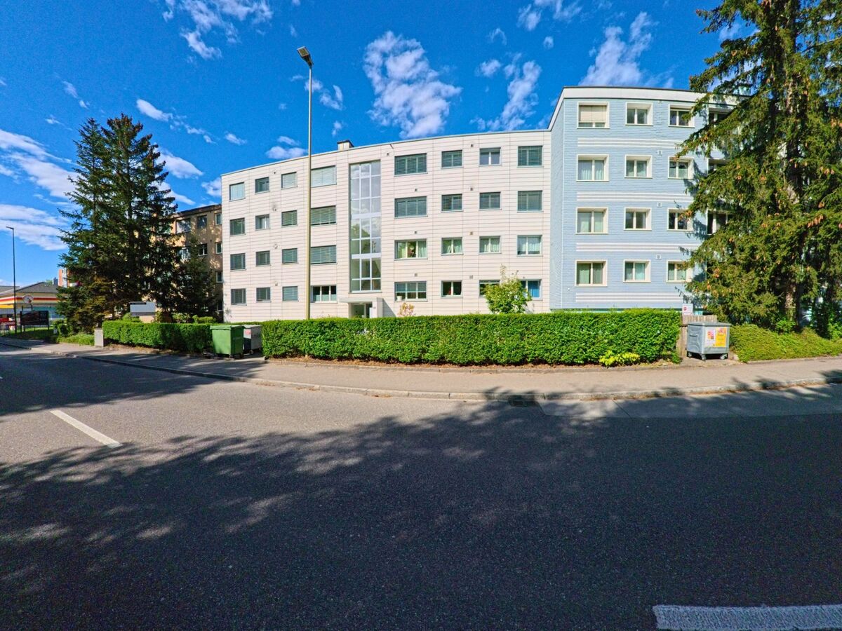 Ein mehrstöckiges Wohnhaus mit heller Fassade steht in Embrach, PLZ 8424 im Kanton Zürich, an einer ruhigen Seitenstrasse neben hohen Bäumen und gepflegten Hecken.