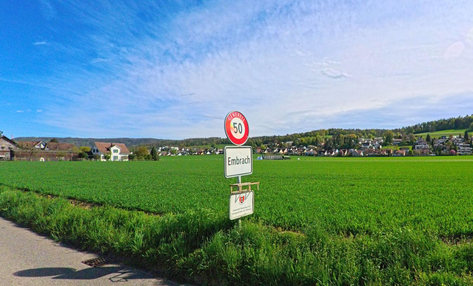 Ein Ortseingangsschild markiert den Übergang nach Embrach, PLZ 8424 im Kanton Zürich, entlang eines Landweges mit Blick auf Felder und die angrenzende Wohnsiedlung.