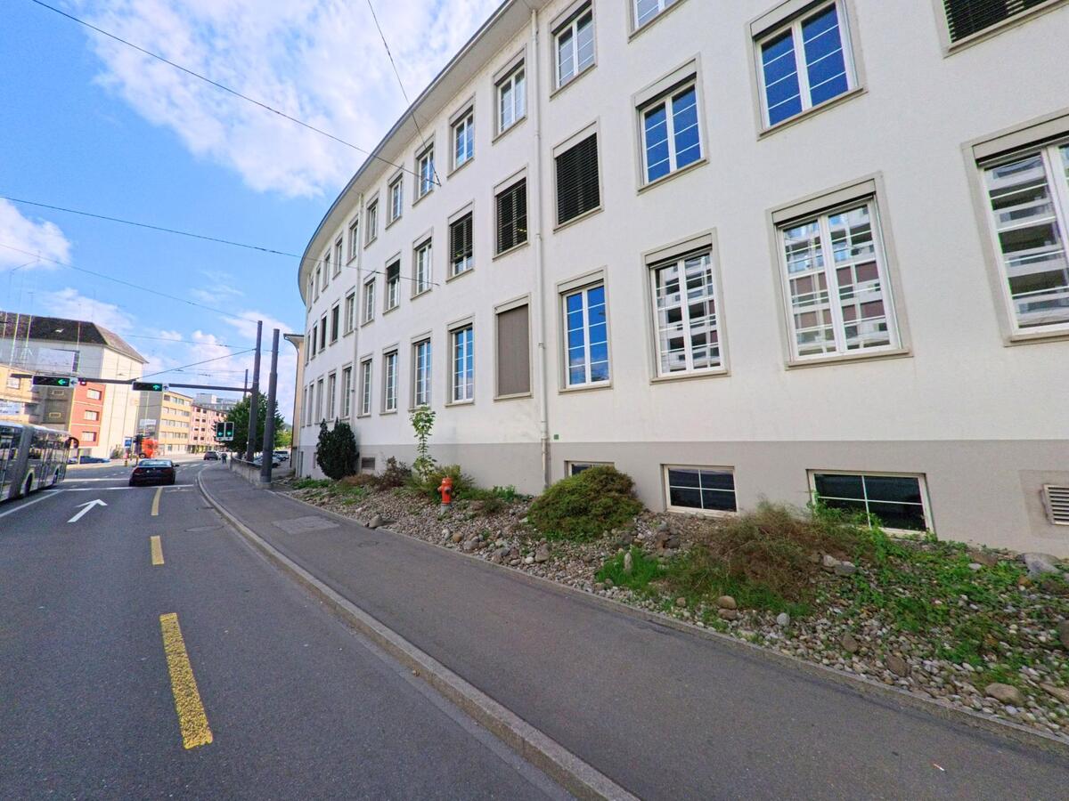 Eine Strasse in Emmen (6020, Kanton Luzern) fuehrt an einem hellen, gebogenen Gebaeude mit vielen Fenstern entlang.